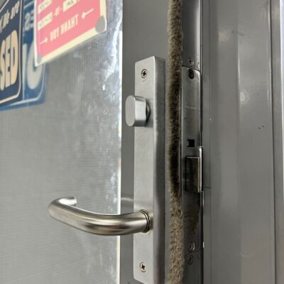 Newly installed stainless steel door lock handle on a metal door with visible business hour signs in Dandenong office.