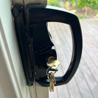 Close-up of a black sliding door lock with keys inserted, mounted on a white door frame with an outdoor view of a wooden deck and greenery.