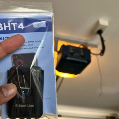 Hand holding a packaged Steel-Line BHT4 garage door remote in front of a garage door opener.