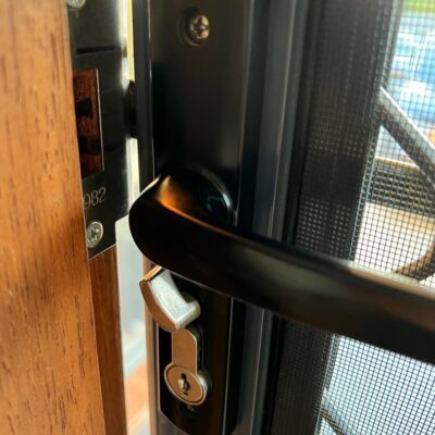 Close-up view of a black metal door lock on a wooden frame with a mesh screen in the background.