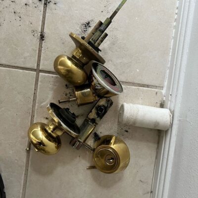 Disassembled brass door lock parts on a tiled floor with black smudges and a white cylindrical object nearby.